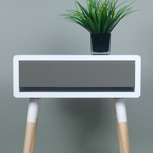 Puede incluir: Mesita de noche blanca con un caj&oacute;n y patas de madera. El caj&oacute;n es gris y las patas est&aacute;n pintadas de blanco con madera natural visible en la parte inferior.