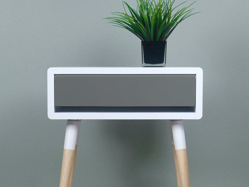 Puede incluir: Mesita de noche blanca con un caj&oacute;n y patas de madera. El caj&oacute;n es gris y las patas est&aacute;n pintadas de blanco con madera natural visible en la parte inferior.
