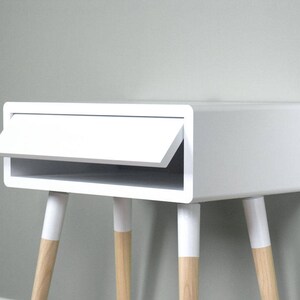 Puede incluir: Mesita de noche blanca con un caj&oacute;n y un estante. La mesa tiene cuatro patas de madera con puntas pintadas de blanco.