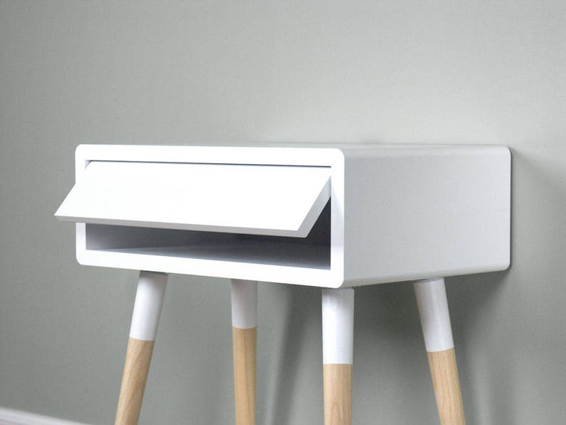 Puede incluir: Mesita de noche blanca con un caj&oacute;n y un estante. La mesa tiene cuatro patas de madera con puntas pintadas de blanco.