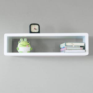 May include: White floating shelf with a green frog plush toy, a black clock, and a stack of books.