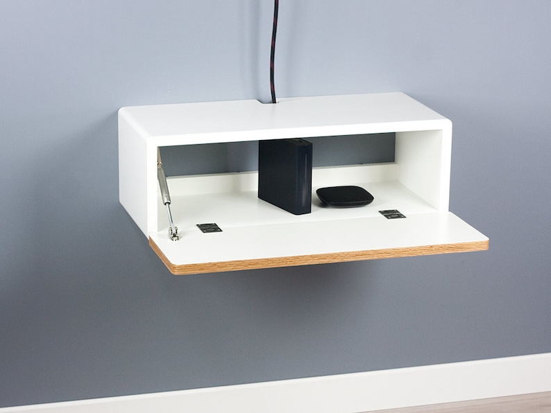 May include: White wall-mounted shelf with a drop-down front. The shelf has a wood trim and is open to reveal a black cable box and a black router.