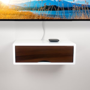 May include: A white floating media console with a dark brown drawer. A small, dark gray remote sits on top. The console is mounted on a white wall, below a television displaying a scenic landscape.