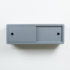 Peut inclure: Un meuble de rangement mural gris rectangulaire avec une porte coulissante. L'unité comporte une zone encastrée et deux trous circulaires noirs. Le design est minimaliste et moderne, adapté à divers besoins de stockage.