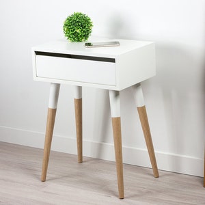 Puede incluir: Mesita de noche blanca con un caj&oacute;n y cuatro patas de madera. Las patas est&aacute;n pintadas de blanco con madera natural en la parte inferior. Una peque&ntilde;a planta verde se encuentra en la parte superior de la mesa.