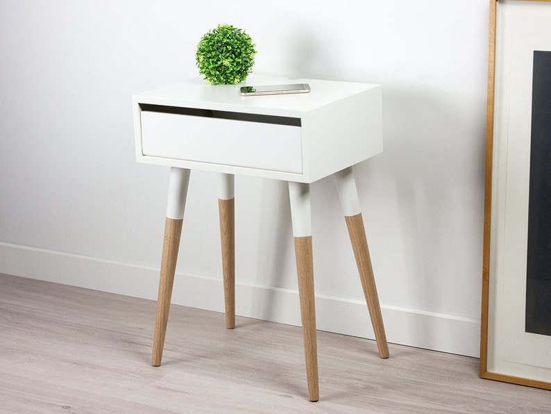 Puede incluir: Mesita de noche blanca con un caj&oacute;n y cuatro patas de madera. Las patas est&aacute;n pintadas de blanco con madera natural en la parte inferior. Una peque&ntilde;a planta verde se encuentra en la parte superior de la mesa.