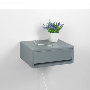 May include: A gray floating nightstand with a drawer and a white cord hanging down. A small potted plant with purple flowers sits on top of the nightstand.