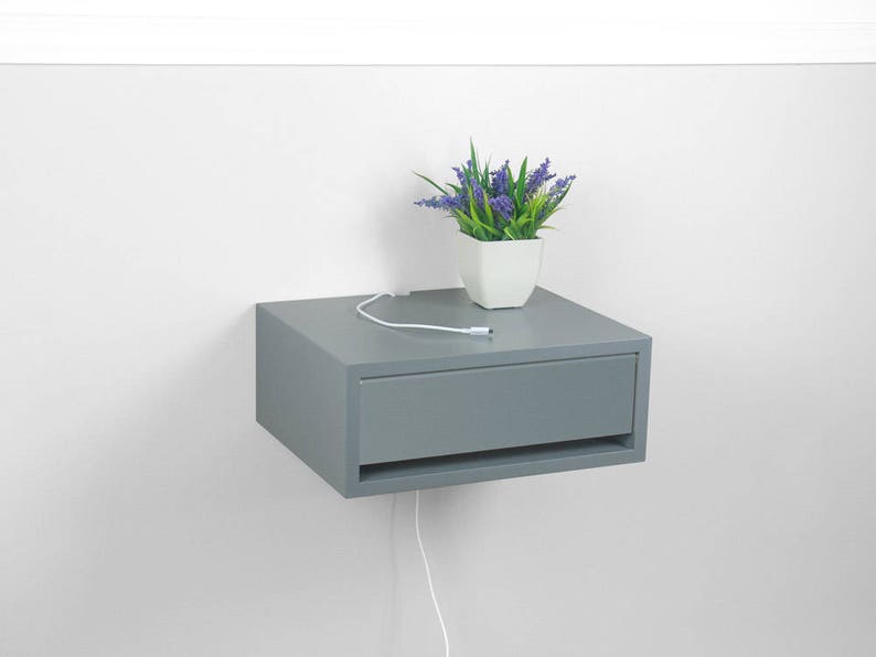 May include: A gray floating nightstand with a drawer and a white cord hanging down. A small potted plant with purple flowers sits on top of the nightstand.