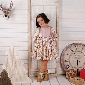 May include: A young girl wearing a yellow and pink floral dress with a pink bodice and a bow on each shoulder. She is standing in front of a white wooden wall with a wooden door behind her. There are two wooden Christmas trees in the foreground.