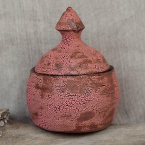 Op de afbeelding: Een handgemaakte keramische pot met deksel, met een getextureerd, gekraakt oppervlak. De pot is voornamelijk een stoffige roze kleur, met donkerbruine accenten. Het deksel is bekroond met een conische knop. Het ontwerp roept een rustieke, ambachtelijke stijl op.