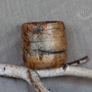 Puede incluir: Una taza de madera con un diseño marmoleado marrón y negro. La taza está sentada en una rama con corteza blanca.