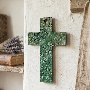 Może przedstawiać: Zielony ceramiczny krzyż z wypukłym, tłoczonym wzorem kwiatowym na całej powierzchni. Krzyż ma błyszczące wykończenie z różnymi odcieniami ciemnej i jasnej zieleni oraz posiada mały okrągły otwór u góry, który służy do zawieszenia.