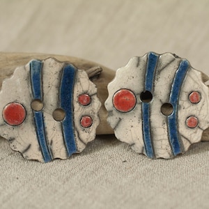Peut inclure: Deux boutons en céramique avec un fond blanc, des rayures bleues et des points rouges. Les boutons ont un bord festonné.