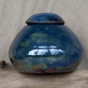 Op de afbeelding: Een blauwe keramische urn met een deksel. De urn heeft een gevlekt blauw en groen glazuur met gouden accenten.