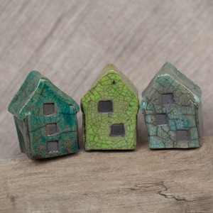 Puede incluir: Tres pequeñas casas de cerámica en tonos verdes y azules con un acabado de esmalte craquelado. Las casas están dispuestas en fila sobre una superficie de madera.