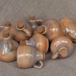 May include: A collection of small, handcrafted ceramic bottles with cork stoppers. The bottles are glazed in a range of brown, beige and white shades, with a speckled finish.