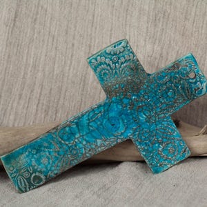 Op de afbeelding: Een turquoiseblauw keramisch kruis met een wit bloemenpatroon. Het kruis heeft een verweerde, gebarsten afwerking.