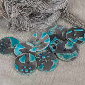 Può includere: Una collezione di bottoni in ceramica fatti a mano in vari design. I bottoni sono principalmente grigi con accenti turchesi e presentano motivi unici. Un fascio di filo grigio è sullo sfondo.