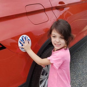 May include: A young person in a pink shirt and shorts touches a round sticker on an orange car. The sticker has a handprint design and the word "Pause". The car is parked on a gravel surface.