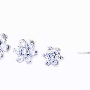 K&ouml;nnte beinhalten: Ein Paar silberne Ohrstecker mit Blumenmuster. Jeder Ohrstecker hat einen Cluster aus kleinen, klaren Steinen in Form einer Blume.