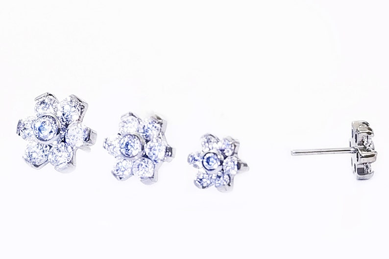 K&ouml;nnte beinhalten: Ein Paar silberne Ohrstecker mit Blumenmuster. Jeder Ohrstecker hat einen Cluster aus kleinen, klaren Steinen in Form einer Blume.