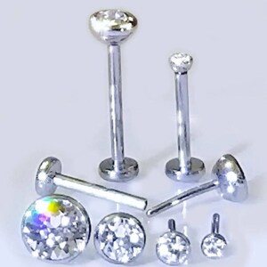 May include: A set of six silver body jewelry pieces with clear crystal accents. The jewelry includes a variety of sizes and styles, including a labret stud, a nose ring, and a lip ring.