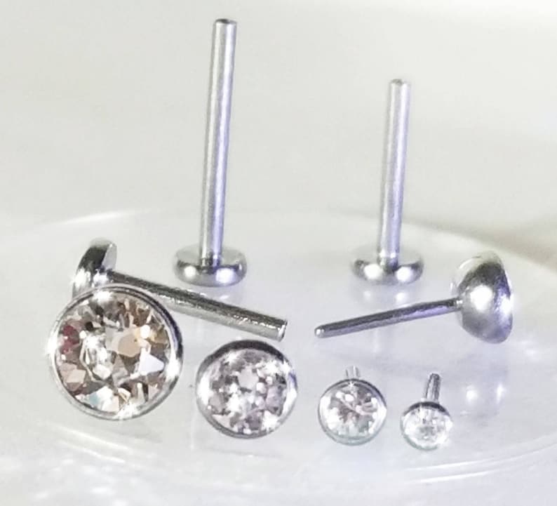 May include: A set of five silver nose studs with clear crystal gems. The studs are different sizes and have a flat back.