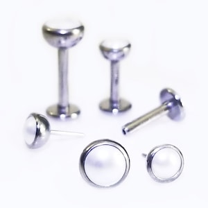 May include: Five silver body jewelry pieces with white pearl-like tops. The pieces are different sizes and styles, including a labret stud, a barbell, and a ring.