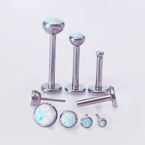 Peut inclure: Un ensemble de sept bijoux de corps argentés avec des pierres ressemblant à des opales. Les pièces sont de différentes tailles et formes, notamment une barre, un labret et un clou.