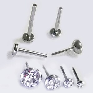 May include: A set of six silver body jewelry pieces. The set includes three labret studs with clear round gems, two labret studs with a smooth round top, and one labret stud with a flat top.