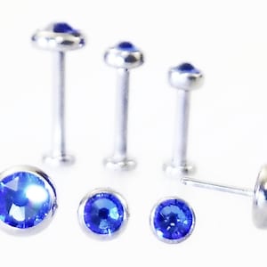 May include: A collection of silver-colored body jewelry, including several flat-backed labret studs and a single earring stud. Each piece features a vibrant, round, blue gemstone set in a bezel setting. The jewelry is displayed against a white background.