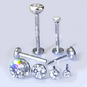 May include: A collection of silver-colored body jewelry, including several labret studs and small, round pendants. The studs have a flat base and a long post, topped with a clear, faceted gemstone. The pendants have a small loop for attachment.