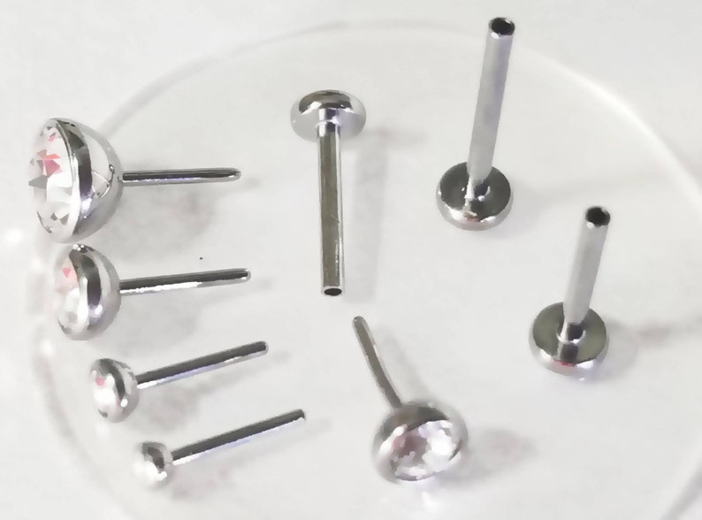 May include: A set of six silver nose rings with clear crystal stones. The rings are different sizes and styles, including a straight post with a ball end, a curved post with a ball end, and a straight post with a crystal stone.