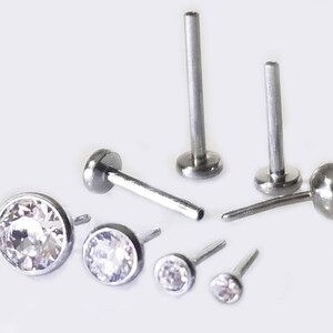 May include: A set of six silver body jewelry pieces with clear crystal accents. The jewelry includes two labret studs, two nose rings, and two small stud earrings.
