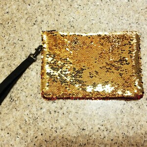 Puede incluir: Clutch rectangular cubierta de lentejuelas doradas con una correa de mu&ntilde;eca negra sujeta por un cierre met&aacute;lico. Las lentejuelas cambian a un color rosa rojizo en el borde inferior. El fondo es una superficie clara y moteada.
