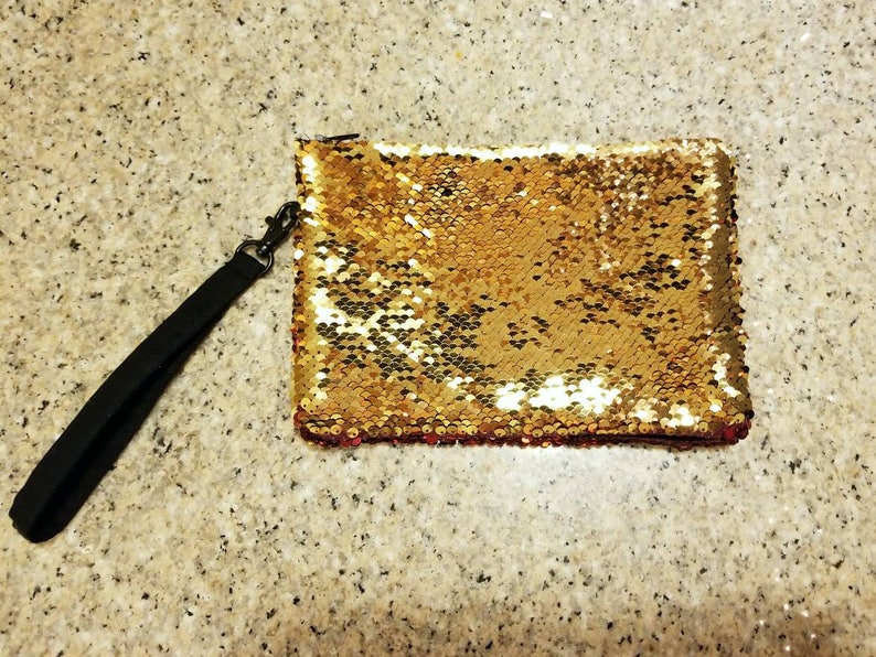 Puede incluir: Clutch rectangular cubierta de lentejuelas doradas con una correa de mu&ntilde;eca negra sujeta por un cierre met&aacute;lico. Las lentejuelas cambian a un color rosa rojizo en el borde inferior. El fondo es una superficie clara y moteada.