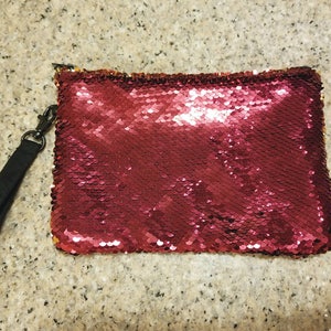 Puede incluir: Un bolso de mano rectangular con lentejuelas rojas, con una correa de mu&ntilde;eca negra sujeta a una cremallera. Las lentejuelas crean un efecto brillante. El bolso de mano est&aacute; dise&ntilde;ado para llevarse en la mano.