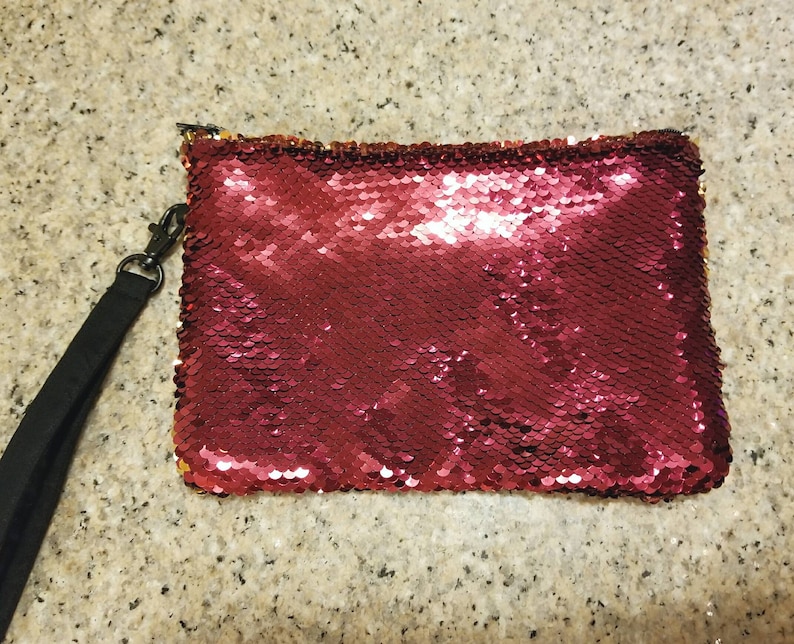 Puede incluir: Un bolso de mano rectangular con lentejuelas rojas, con una correa de mu&ntilde;eca negra sujeta a una cremallera. Las lentejuelas crean un efecto brillante. El bolso de mano est&aacute; dise&ntilde;ado para llevarse en la mano.