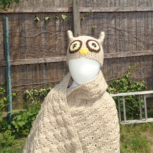 以下が含まれることがあります： 茶色と黄色のアクセントが付いた、ベージュのかぎ針編みのフクロウの帽子と、お揃いのスカーフ。 帽子には大きくて丸い目と小さな耳のような突起があります。 スカーフはマネキンに掛けられています。