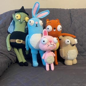 May include: A group of five handmade crochet stuffed animals. The collection includes a green witch, a blue bunny, an orange cat, a pink bunny, and a tan bear. Each has large, expressive eyes and unique features.