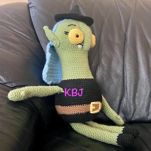 Könnte beinhalten: Eine grüne Häkel-Hexenpuppe mit einem schwarzen Hut, blauen Haaren und einem schwarzen und braunen Gürtel. Die Puppe sitzt auf einem schwarzen Ledersofa. Die Puppe hat ein großes gelbes Auge und ein breites Grinsen. Die Puppe ist mit "KBJ" beschriftet.