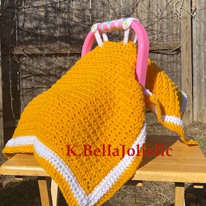 Puede incluir: Una manta de bebé de ganchillo amarillo mostaza con un borde blanco, sobre un asa rosa para el portabebés. La manta está hecha a mano y tiene un aspecto texturizado. El texto "K.BellaJolle" es visible.