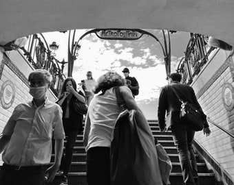 May include: Black and white photograph of people ascending and descending a staircase under an archway. The archway reads "METROPOLITAIN". Some people are wearing face masks. The sky is visible above the archway.