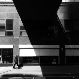 May include: A black and white photo of a person walking in front of a building with large windows. The building has a modern design with a flat roof and a large overhang.