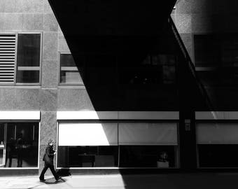 May include: A black and white photo of a person walking in front of a building with large windows. The building has a modern design with a flat roof and a large overhang.