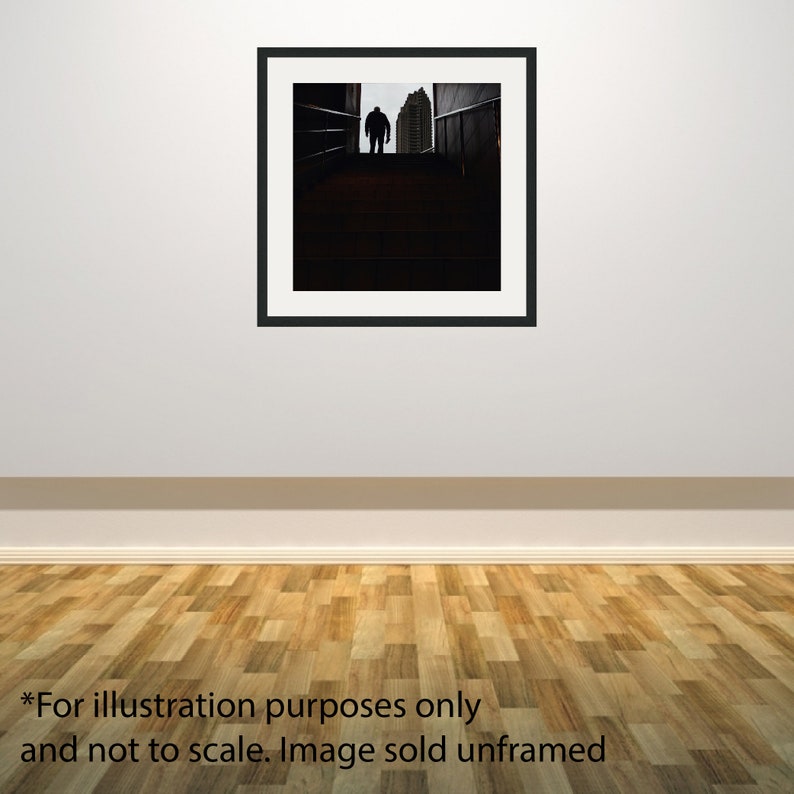 Puede incluir: Fotograf&iacute;a en blanco y negro enmarcada de una persona silueteada en la parte superior de una escalera, con un edificio visible en el fondo. La imagen est&aacute; montada en un marco negro y colgada en una pared blanca.