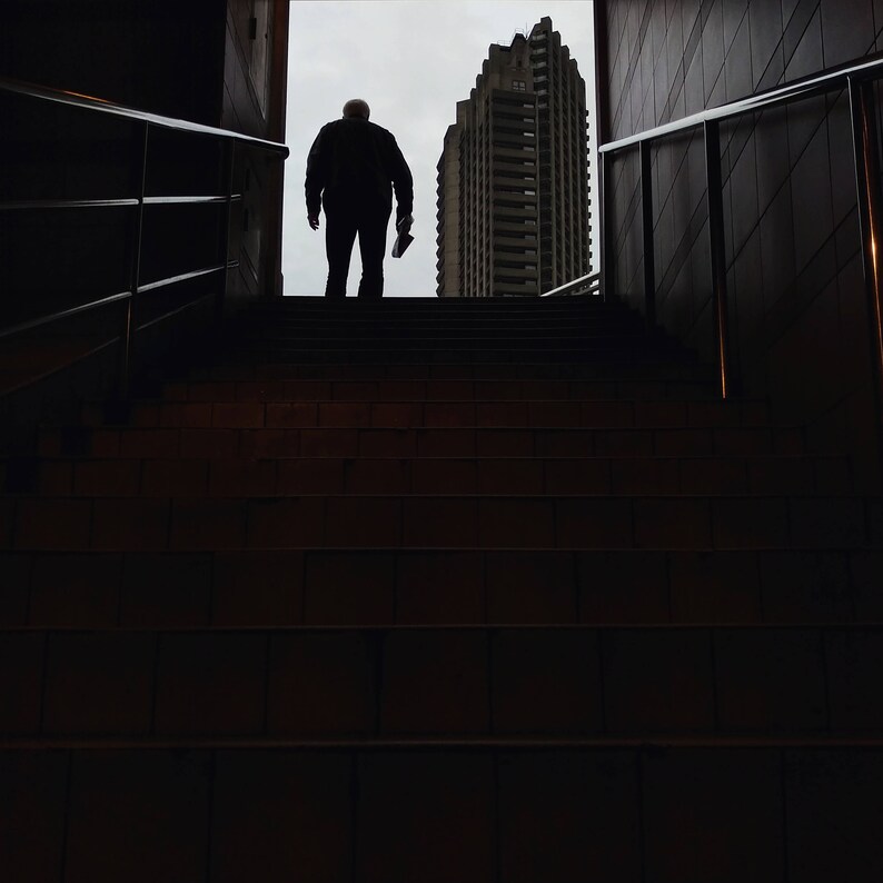 Puede incluir: Silueta de una persona en la parte superior de una escalera, sosteniendo un objeto, con un edificio alto al fondo. La escalera es oscura, que conduce a un cielo brillante y nublado. El edificio es una estructura moderna de varios pisos.