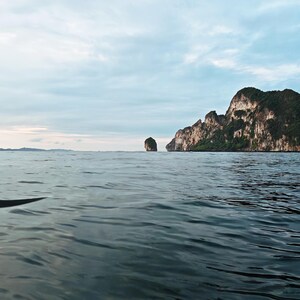 Può includere: Un sereno paesaggio marino con acqua blu scuro calma sotto un cielo nuvoloso. In lontananza, sono visibili una piccola isola e una grande scogliera rocciosa. La scena evoca un senso di tranquillità e bellezza naturale.