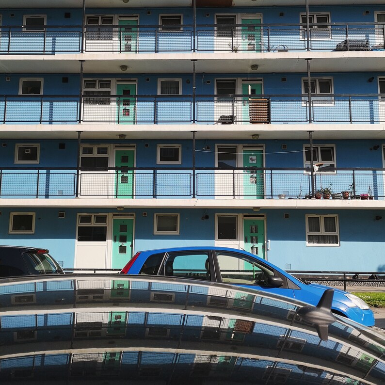 K&ouml;nnte beinhalten: Ein blaues Auto parkt vor einem mehrst&ouml;ckigen Apartmenthaus mit wei&szlig;en Gel&auml;ndern und Fenstern. Das Geb&auml;ude hat eine hellblaue Fassade.
