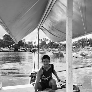 Może przedstawiać: Czarno-białe zdjęcie uśmiechniętej osoby siedzącej w małej łodzi. Łódź znajduje się na wodzie, z baldachimem nad nią. W tle widać linię brzegową z budynkami i palmami.